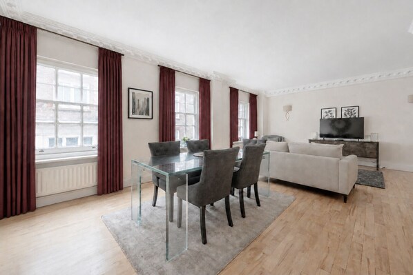 A modern living room with a glass dining table, grey sofas, and a flat-screen TV.