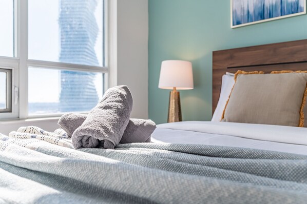 Una cama bien hecha con una colcha blanca y almohadas, una lámpara en una mesita de noche, y vistas de un paisaje urbano a través de una ventana.