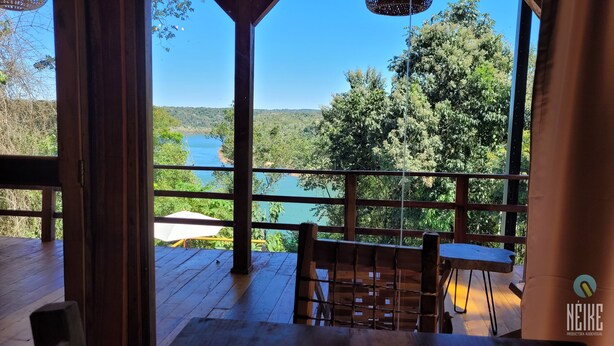 Terraza de madera con mesa y sillas, con vista a un lago y colinas boscosas.