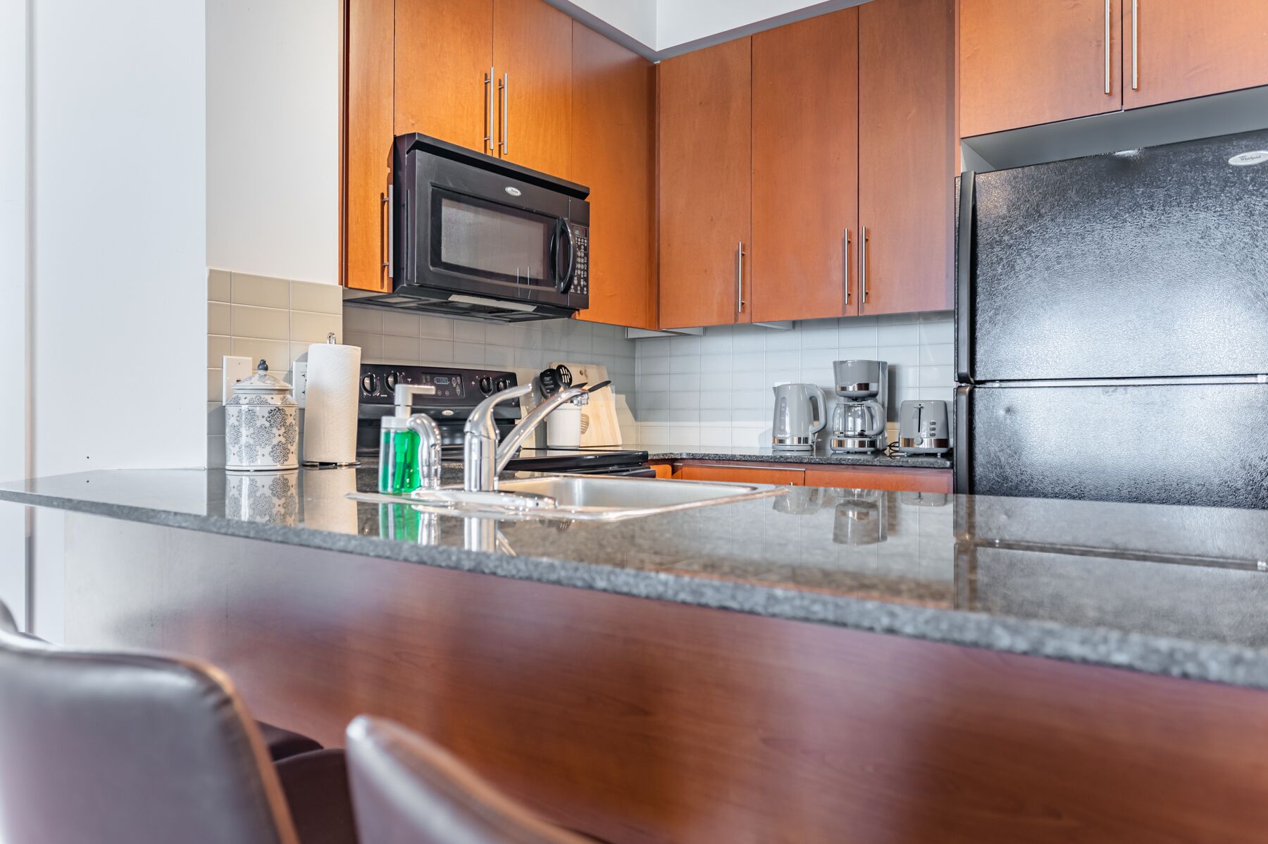 A modern kitchen with wooden cabinets, a black microwave, a black refrigerator, a sink, and a coffee maker.
