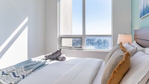 Une chambre d’hôtel avec une grande fenêtre, un lit avec du linge de maison blanc, un plaid à motifs et une vue sur la ville.