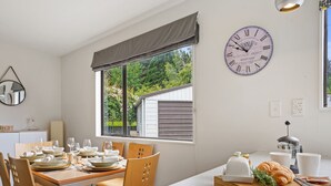 A dining area with a table set for four, a clock on the wall, and a window with a view of greenery. - Rose Cottage - Lake Coleridge Holiday Home (Lake Coleridge)