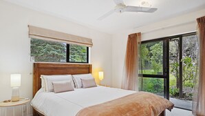 A bedroom with a large bed, a bedside table with a lamp, a window with a view of greenery, and a ceiling fan. - Rose Cottage - Lake Coleridge Holiday Home (Lake Coleridge)