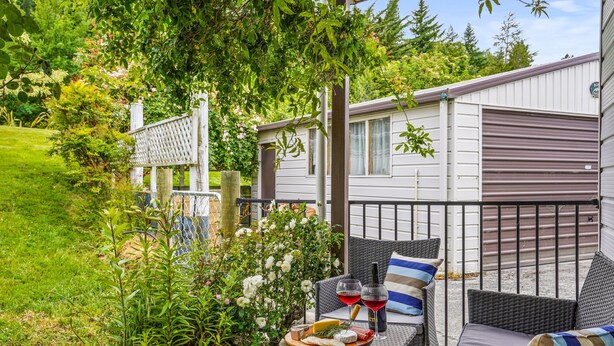 A patio with wicker furniture, a table with a cheese platter and wine glasses, and a view of a mobile home in the background. - Rose Cottage - Lake Coleridge Holiday Home (Lake Coleridge)