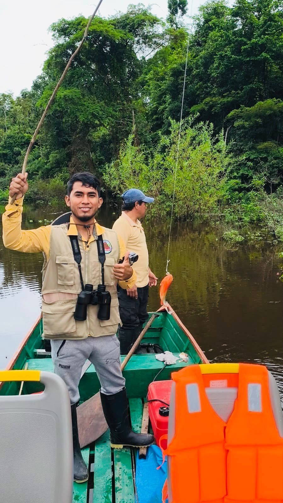 Two individuals in a green boat with an orange life jacket, holding fishing rods, surrounded by dense greenery and a calm river.