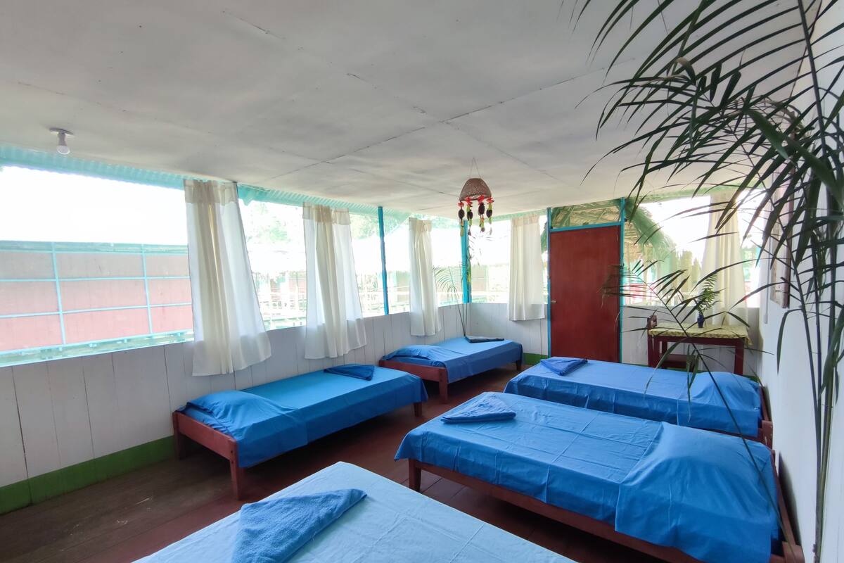 Una habitación con cuatro camas individuales, cada una con cubrecama azul, un banco de madera, una mesita pequeña y una lámpara colgante.