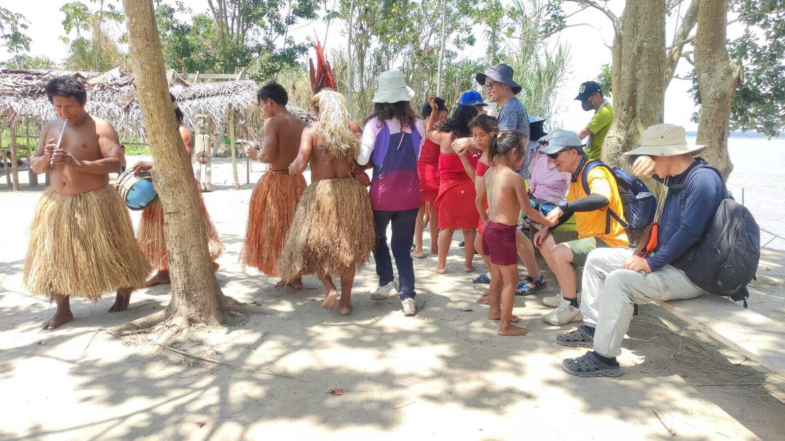 A group of people in traditional attire and casual clothing near a beach, with trees and a thatched structure in the background.