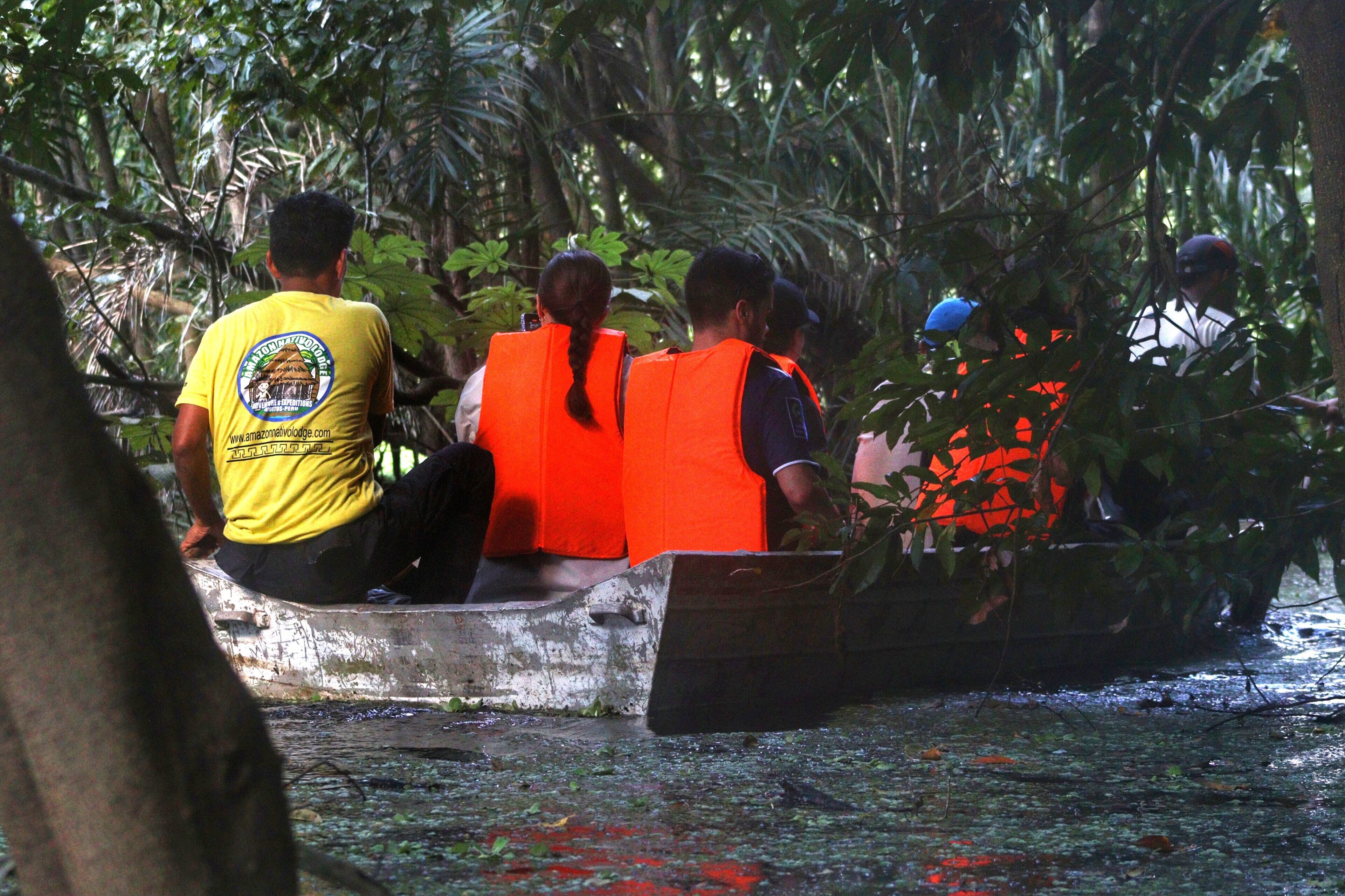 A group of people in a boat wearing life jackets, surrounded by dense foliage.