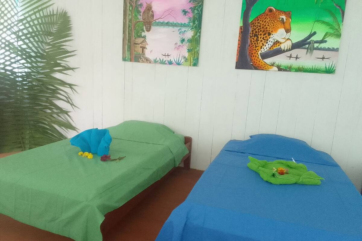 Dos camas individuales con ropa de cama verde y azul, una planta grande en maceta y dos cuadros en la pared.