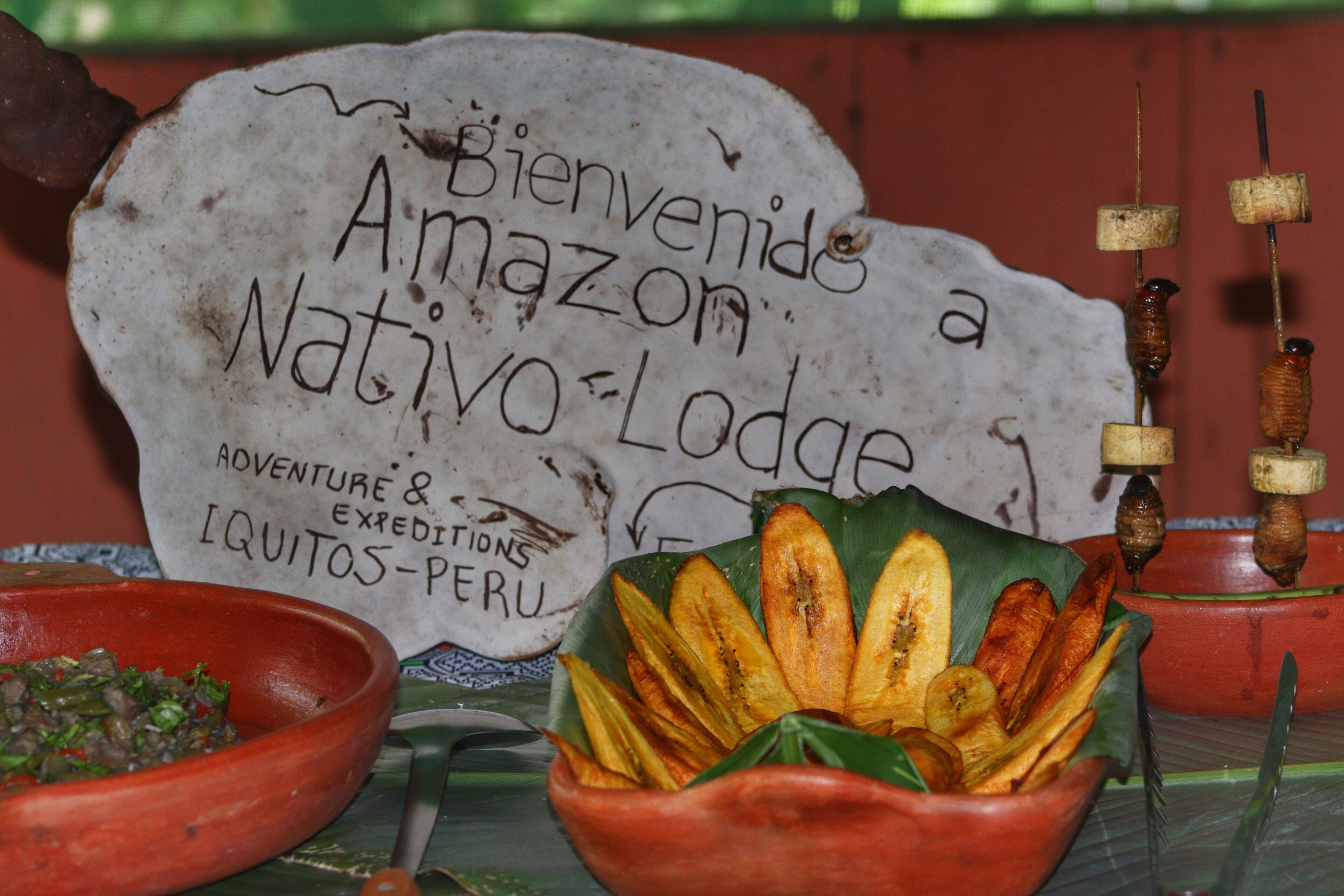 Une pancarte d’accueil pour l’Amazon Native Lodge, une pancarte au style rustique, un bol de nourriture et une présentation de brochettes.