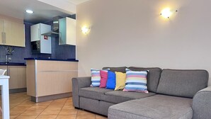 En moderne stue med en grå hjørnesofa, et lille hvidt bord og et tekøkken med mikrobølgeovn og køleskab.