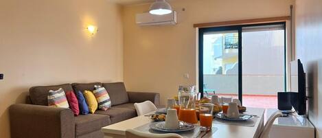 A living room with a sofa, a dining table with breakfast items, a TV, and a balcony with a view.