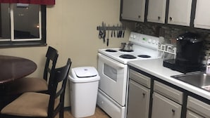 A compact kitchen with a stove, sink, and cabinets. There is a clock on the wall and a window with a red curtain.