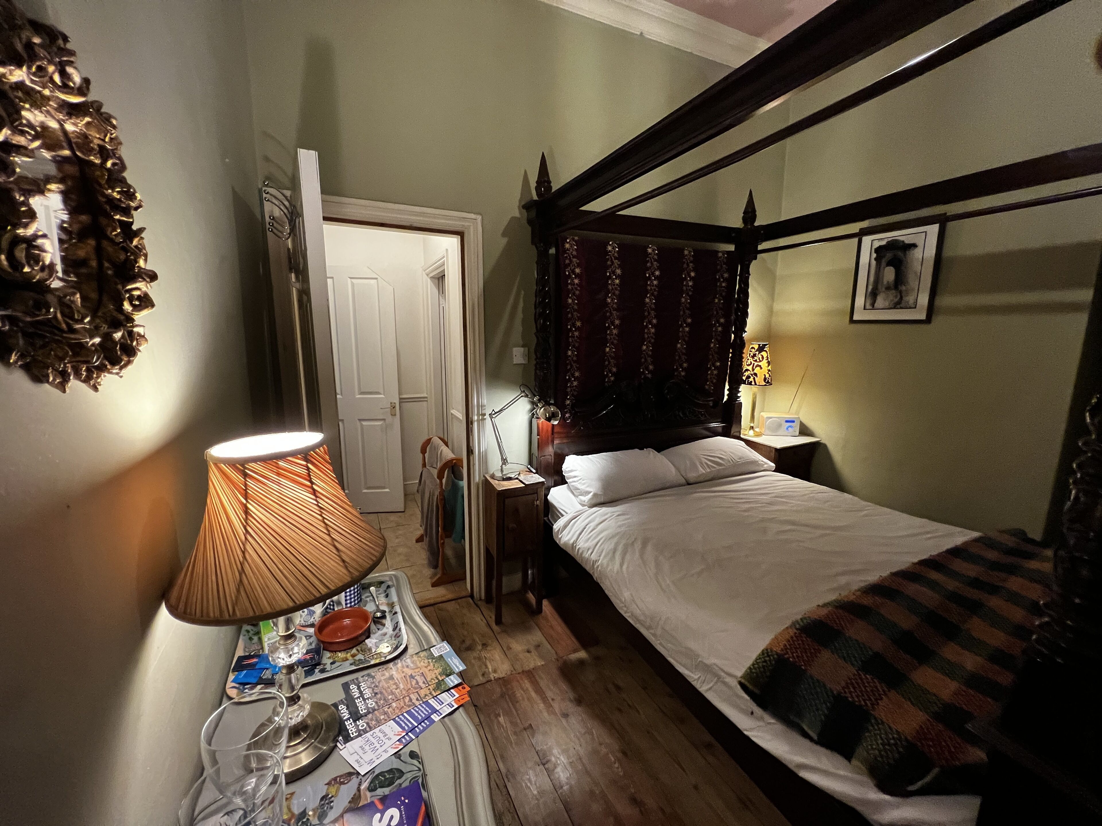 A bedroom with a four-poster bed, a bedside table with a lamp, a small table with magazines and a glass, and a door leading to another room.