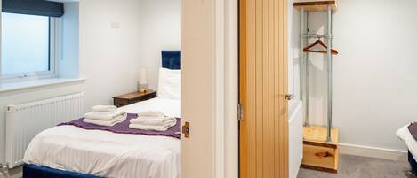 A bedroom with a bed, a bedside table, a window, a radiator, a wooden wardrobe, and a hanging wardrobe.