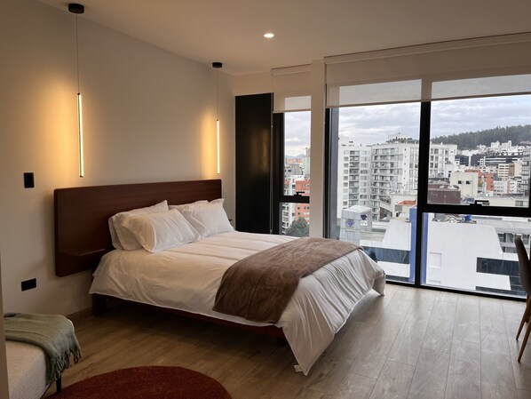 Ein modernes Hotelzimmer mit einem großen Bett und Blick auf die Stadt durch raumhohe Fenster. Außerhalb ist eine Stadtansicht zu sehen.