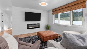 A modern living room with a flat-screen TV mounted on the wall, a fireplace below it, and a large window with a view of greenery. - Fireplace|Garage|Pets|King Bed! (Green Bay)