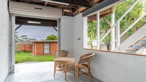 Un espace extérieur aménagé avec une zone salon couverte, comprenant deux fauteuils en osier et une petite table ronde, un mur blanc et une vue sur une cour arrière avec une maison en briques.