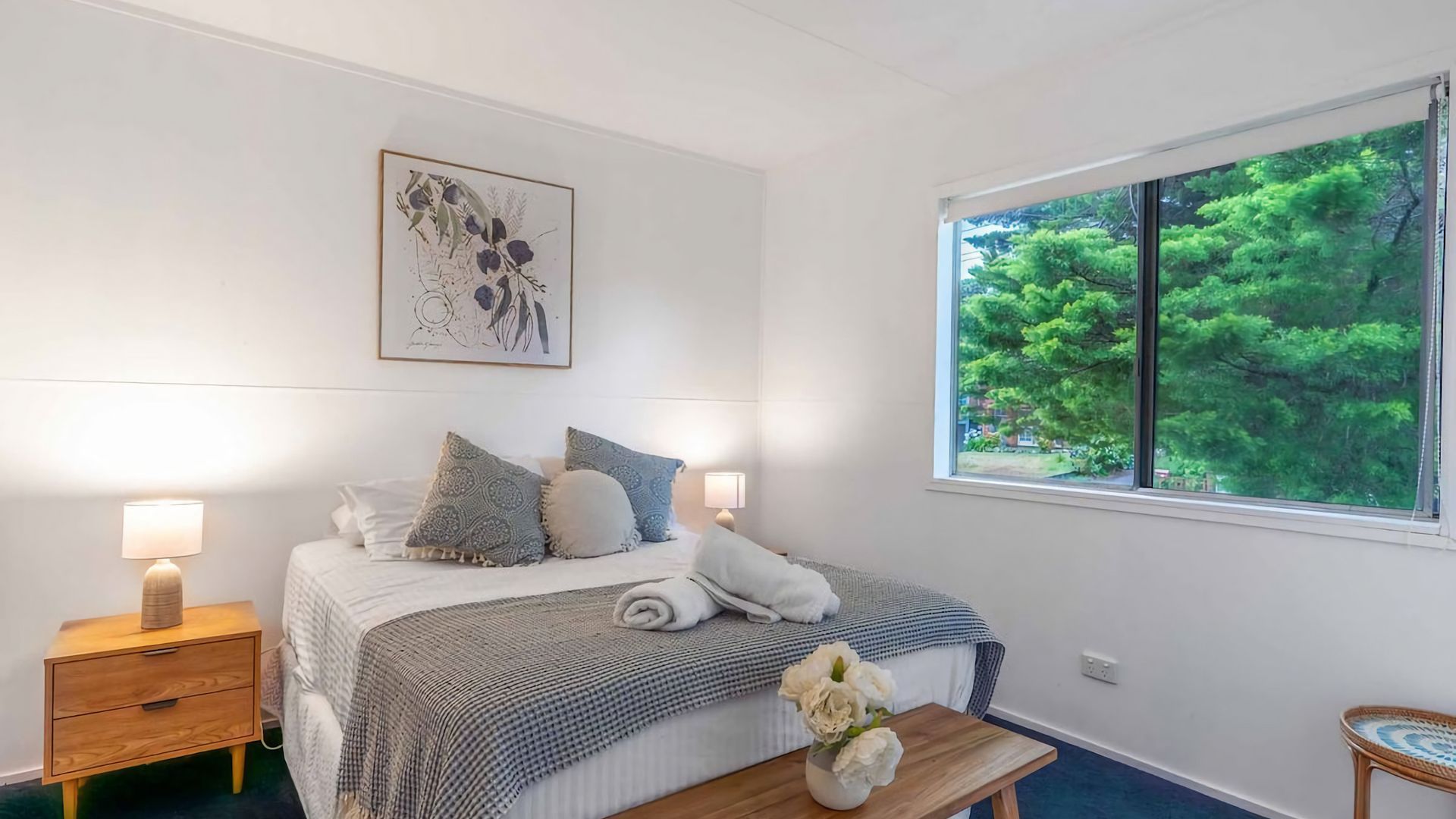 A bedroom with a bed, bedside lamps, a window with a view of trees, and a wall-mounted artwork.