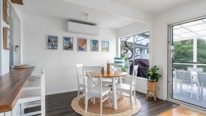 A dining area with a round table, white chairs, and a small side table with a candle. There are framed pictures on the wall and a window with a view of the outdoors. - The Beach Box is a pet friendly rental (Manyana)