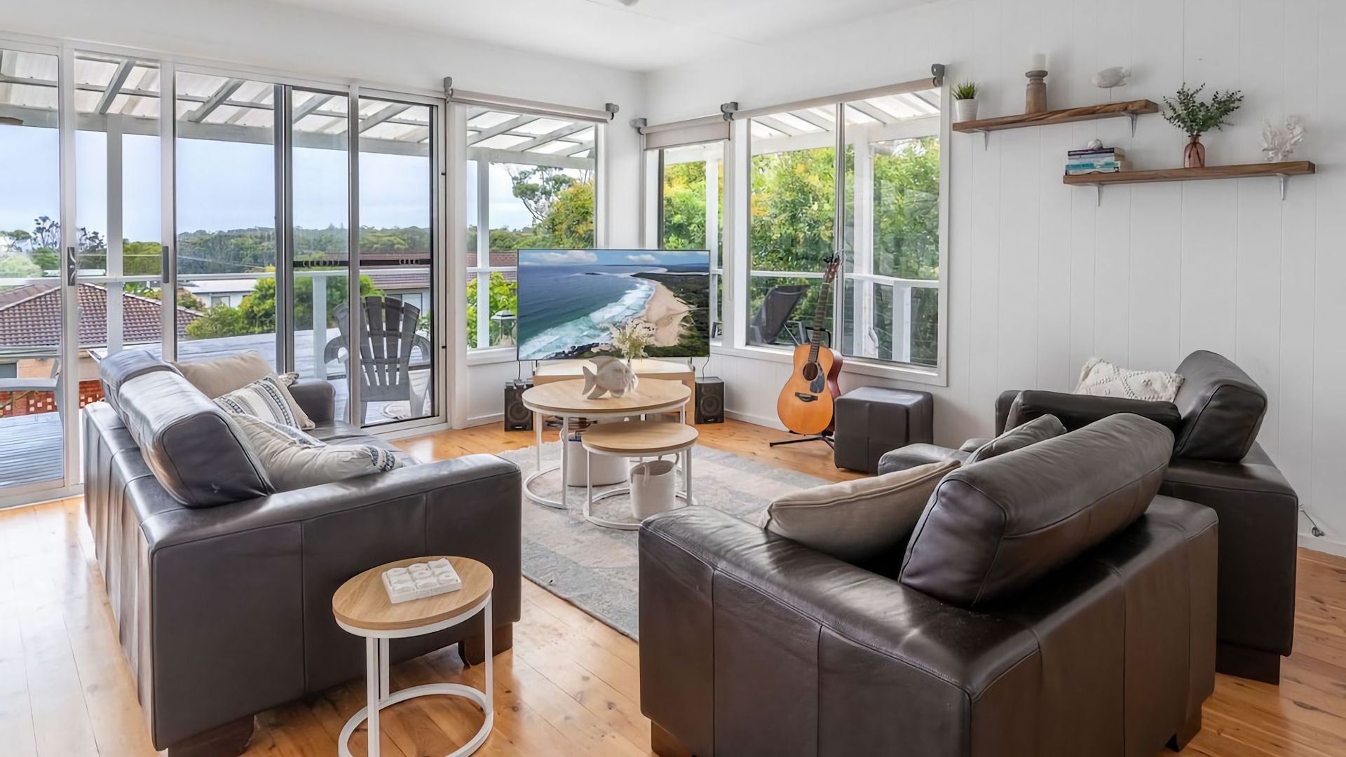 Ein geräumiges Wohnzimmer mit einem großen Fernseher, Ledersofas, einer Gitarre und Blick ins Freie.