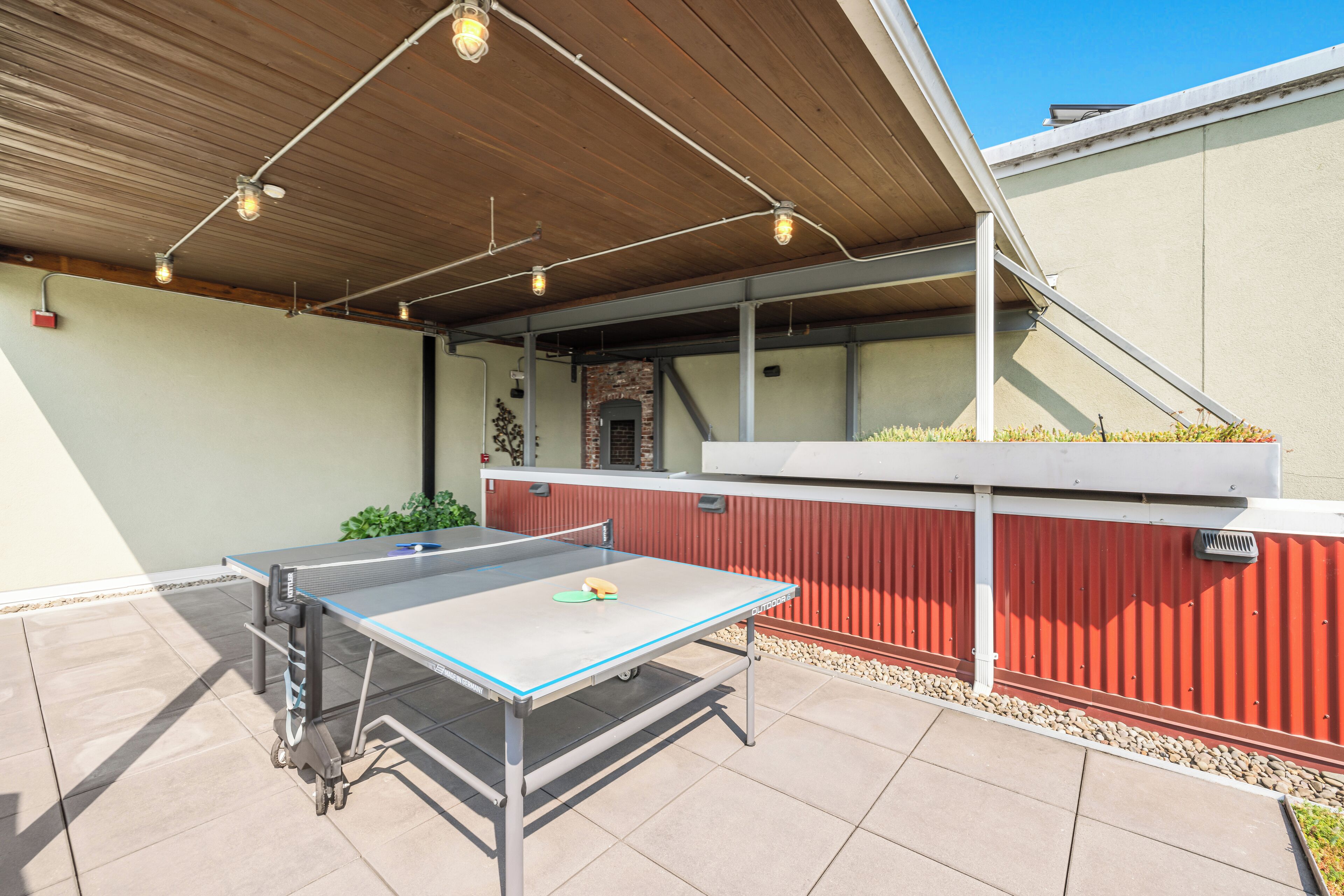 Une table de ping-pong installée à l’extérieur, sous une pergola en bois, avec un mur rouge et une cheminée en briques en arrière-plan.