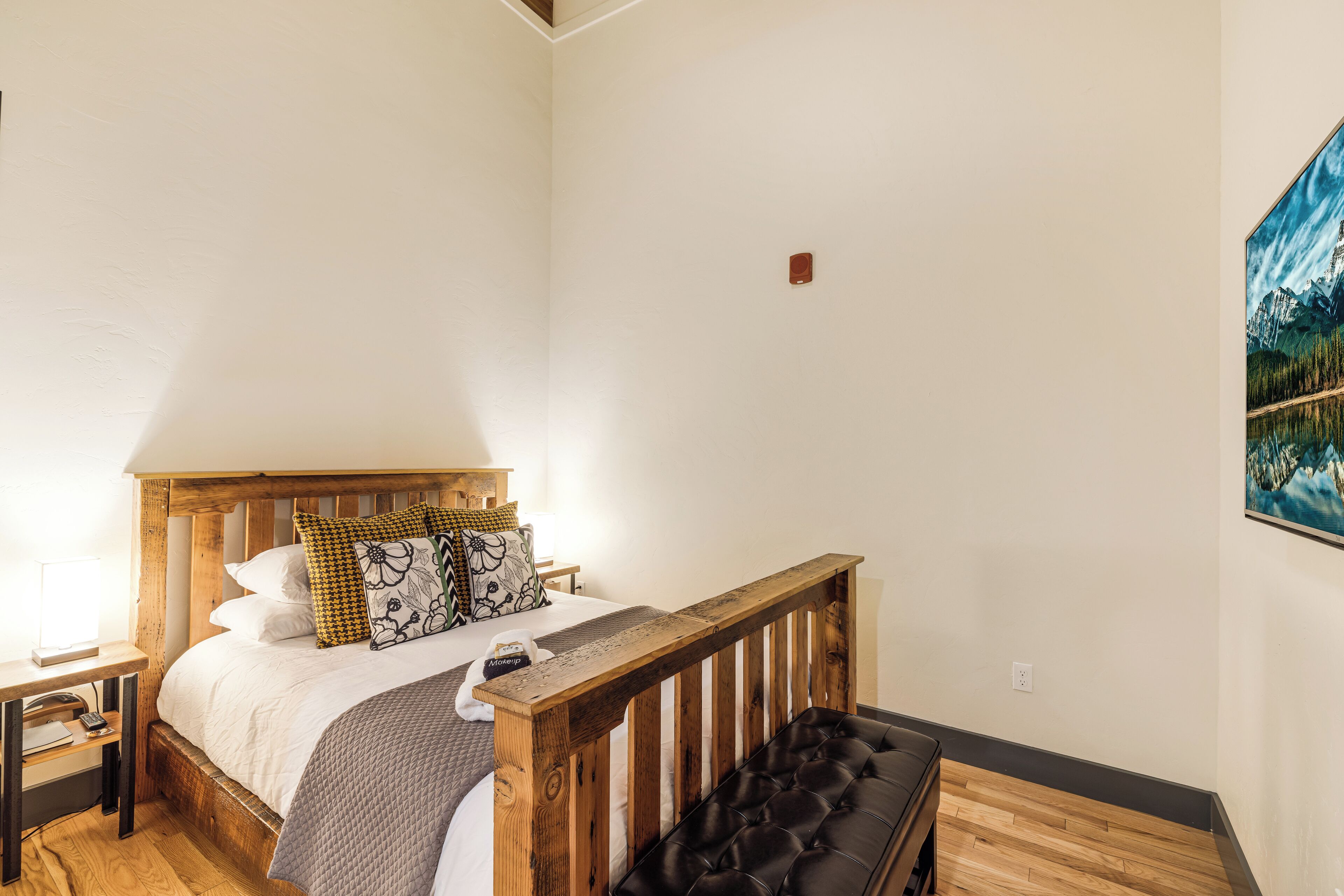 A bedroom with a wooden headboard, a bed with white bedding, a black leather bench, and a painting on the wall.