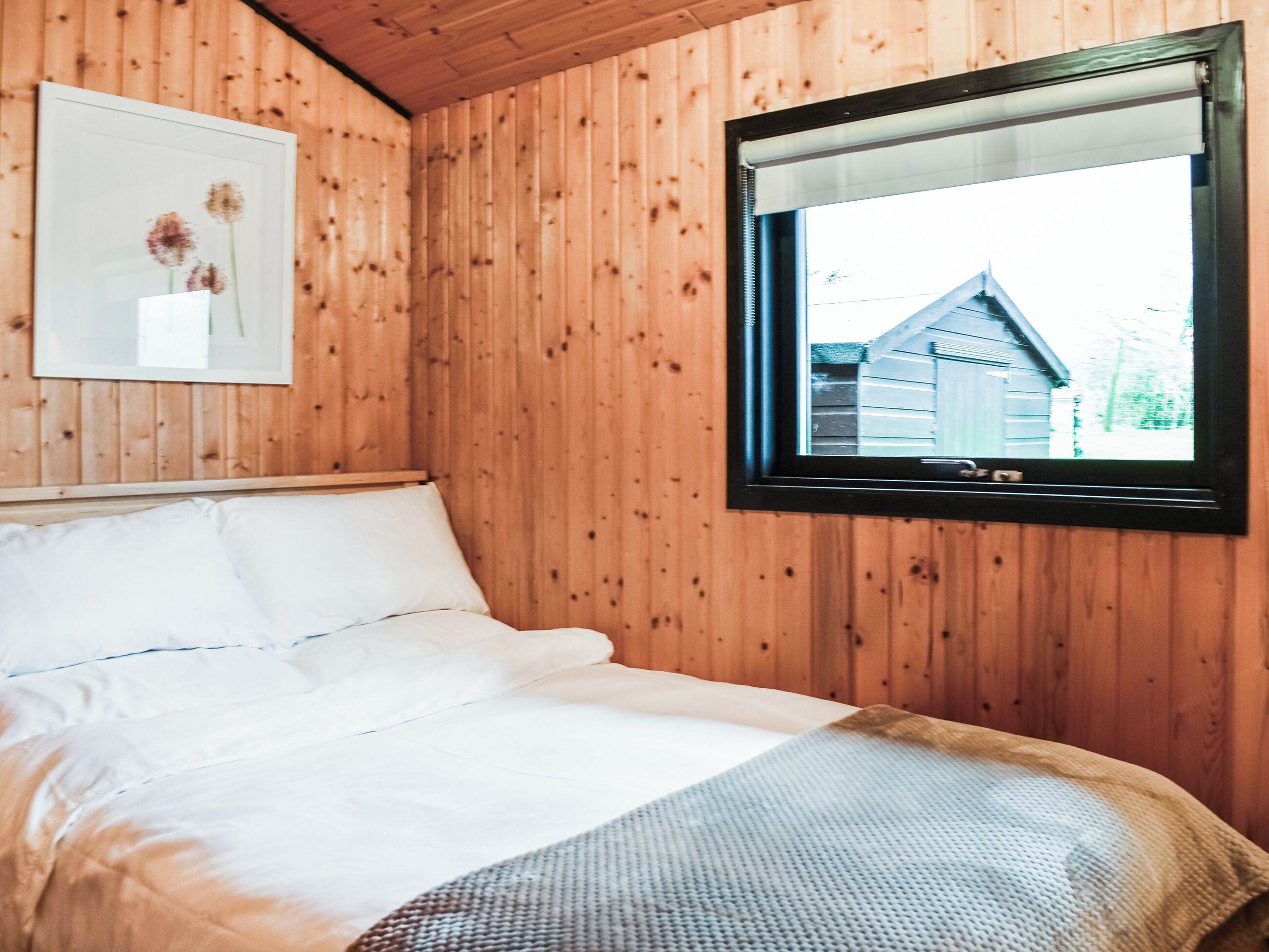 A wooden-paneled room with a bed, a framed picture on the wall, and a window with a view of a house.