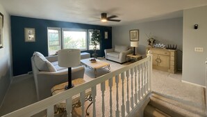 A living room with a white railing, a ceiling fan, and a view of the outdoors through large windows.