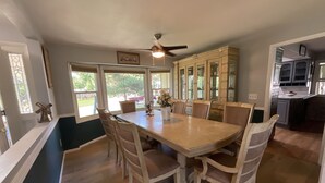 A dining room with a large wooden table, six chairs, a ceiling fan, and a view of the outdoors through large windows.