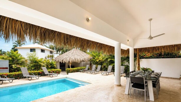 Un área junto a la piscina con sillones de descanso, una mesa de comedor para dos personas y una sombrilla de paja.