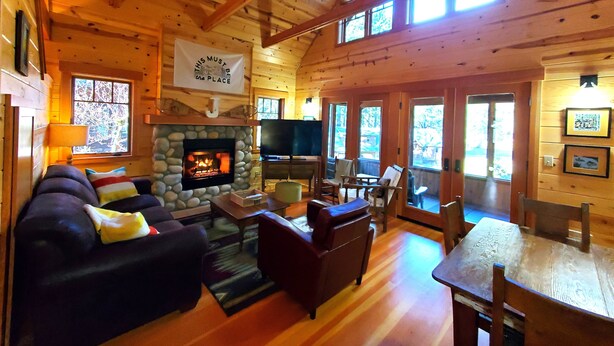 Un intérieur de chalet chaleureux avec une cheminée en pierre, des murs en bois, un coin salon confortable et de grandes fenêtres.