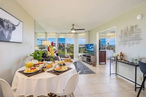 A dining area with a table set for breakfast, a TV in the background, and a view of the landscape. - Oceanview Retreat | Walk to Beach & Restaurants (Wailea-Makena)