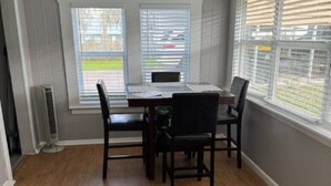 Un comedor con una mesa de madera y cuatro sillas, ventanas amplias con persianas y un ventilador de techo.