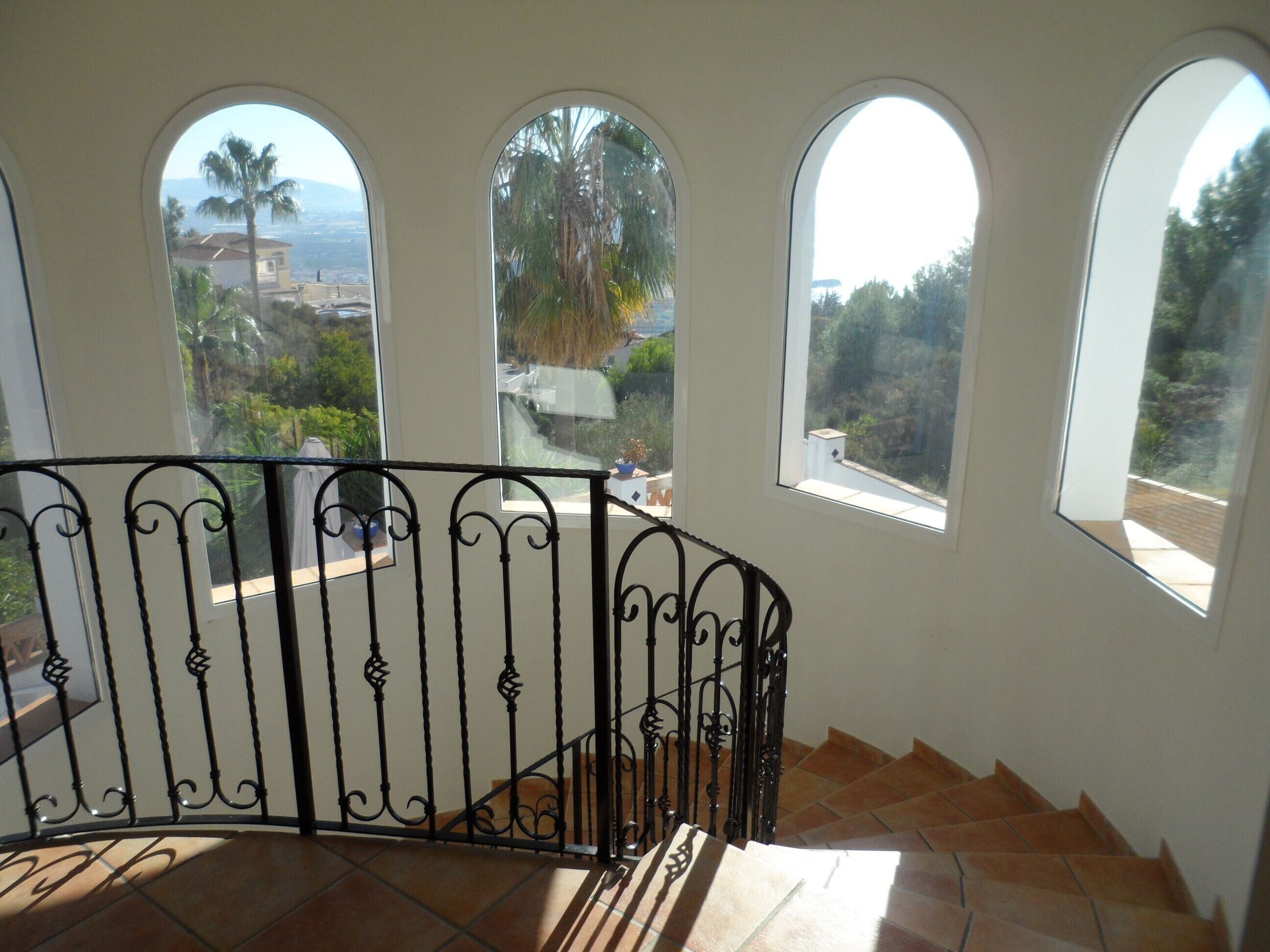 Una scala con ringhiera in ferro battuto che conduce a un balcone con tre finestre ad arco affacciato su un paesaggio con palme e una vista lontana di un orizzonte urbano.