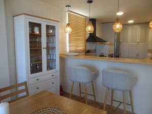 Una cocina con una mesa de madera, dos taburetes de bar, un armario con puertas de vidrio y lámparas colgantes.