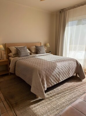 A bedroom with a wooden bed, multiple pillows, a bedside table with a lamp, and a large window with curtains.