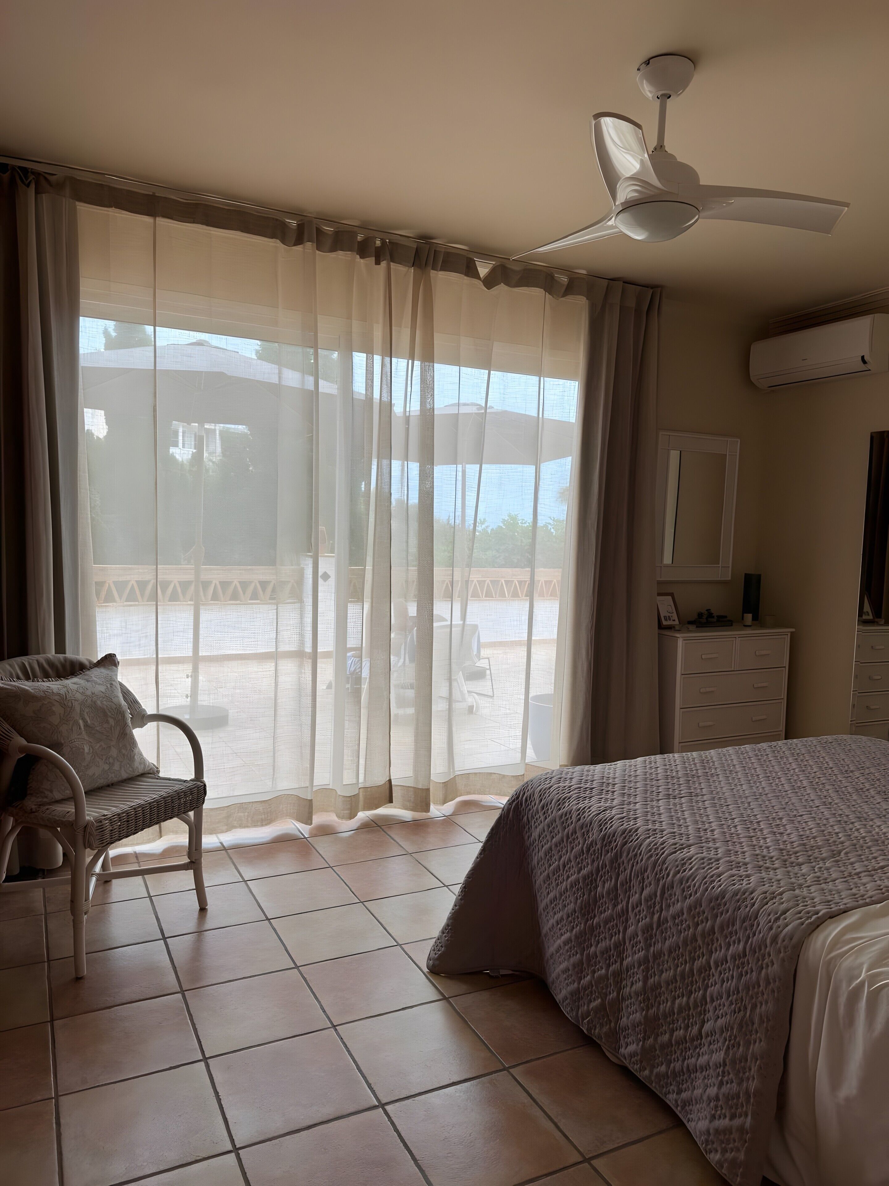 Une chambre à coucher comprenant un lit, une chaise, un ventilateur de plafond, une fenêtre avec des rideaux et un appareil de climatisation.