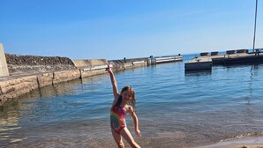 A child playing with a ball on a sandy beach near a body of water with a dock and a flag in the background. - The Beachcomber at Dockside Resort - Near Bohemia and Trails (Lake Linden)