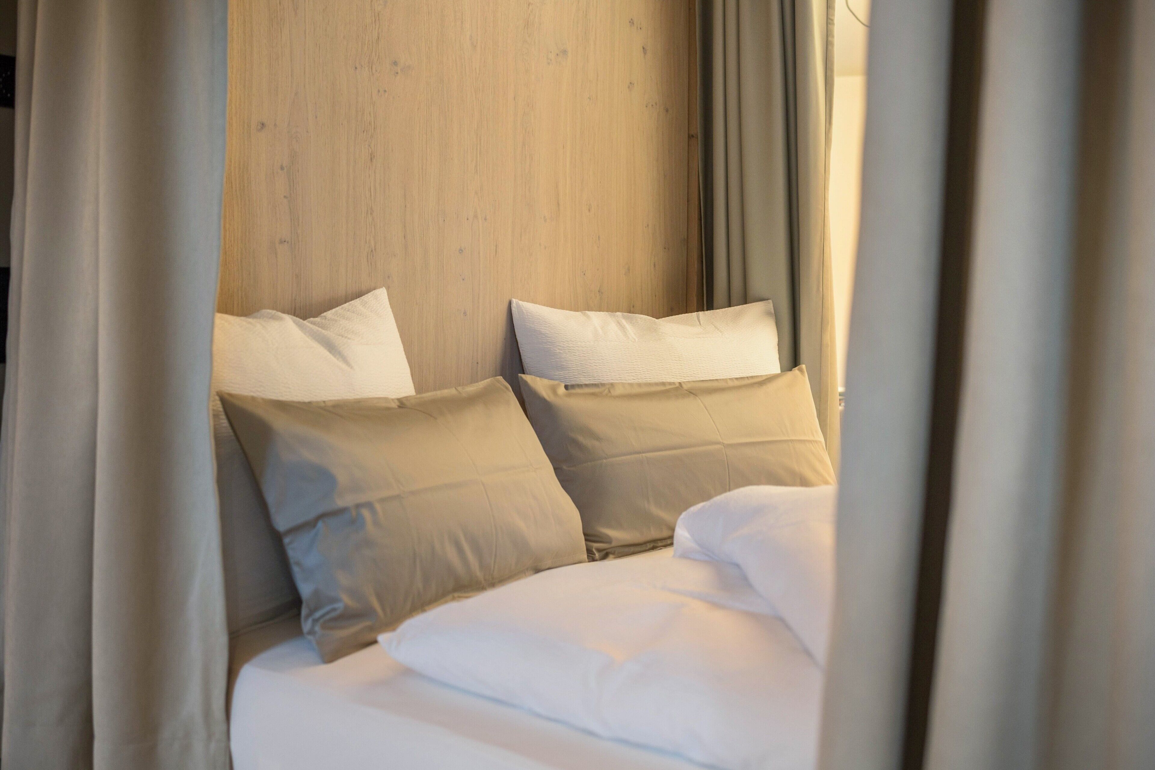 A bed with white bedding and beige pillows, a wooden headboard, and curtains partially drawn.