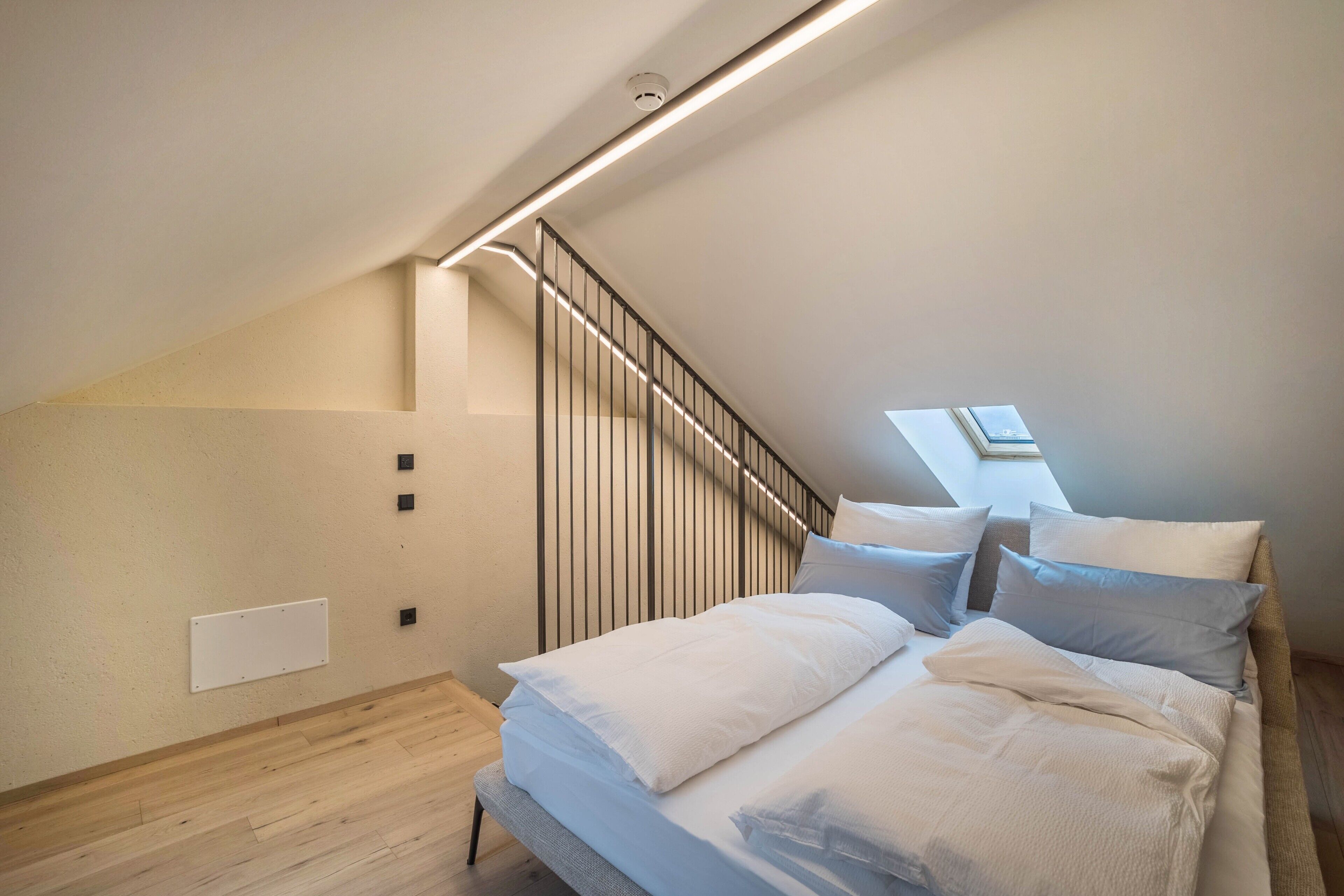 Ein Schlafzimmer mit schrägverdeckter Decke, einem Bett mit weißer Bettwäsche, Holzboden, einer wandmontierten Lampe und einem Oberlicht.