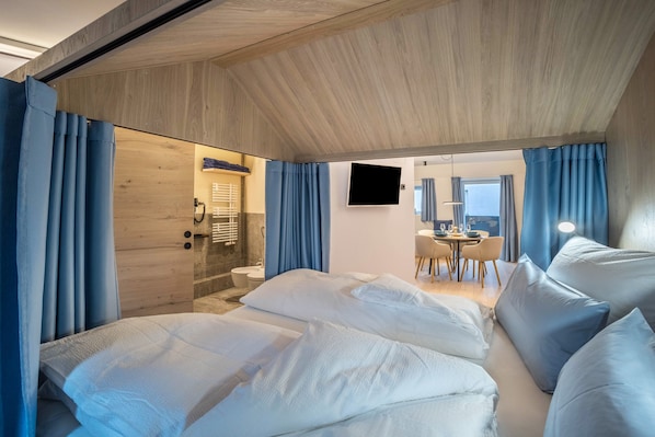 A modern hotel room with a wooden ceiling, a large bed with white linens, a bathroom with a glass shower enclosure, and a dining area with a table and chairs.