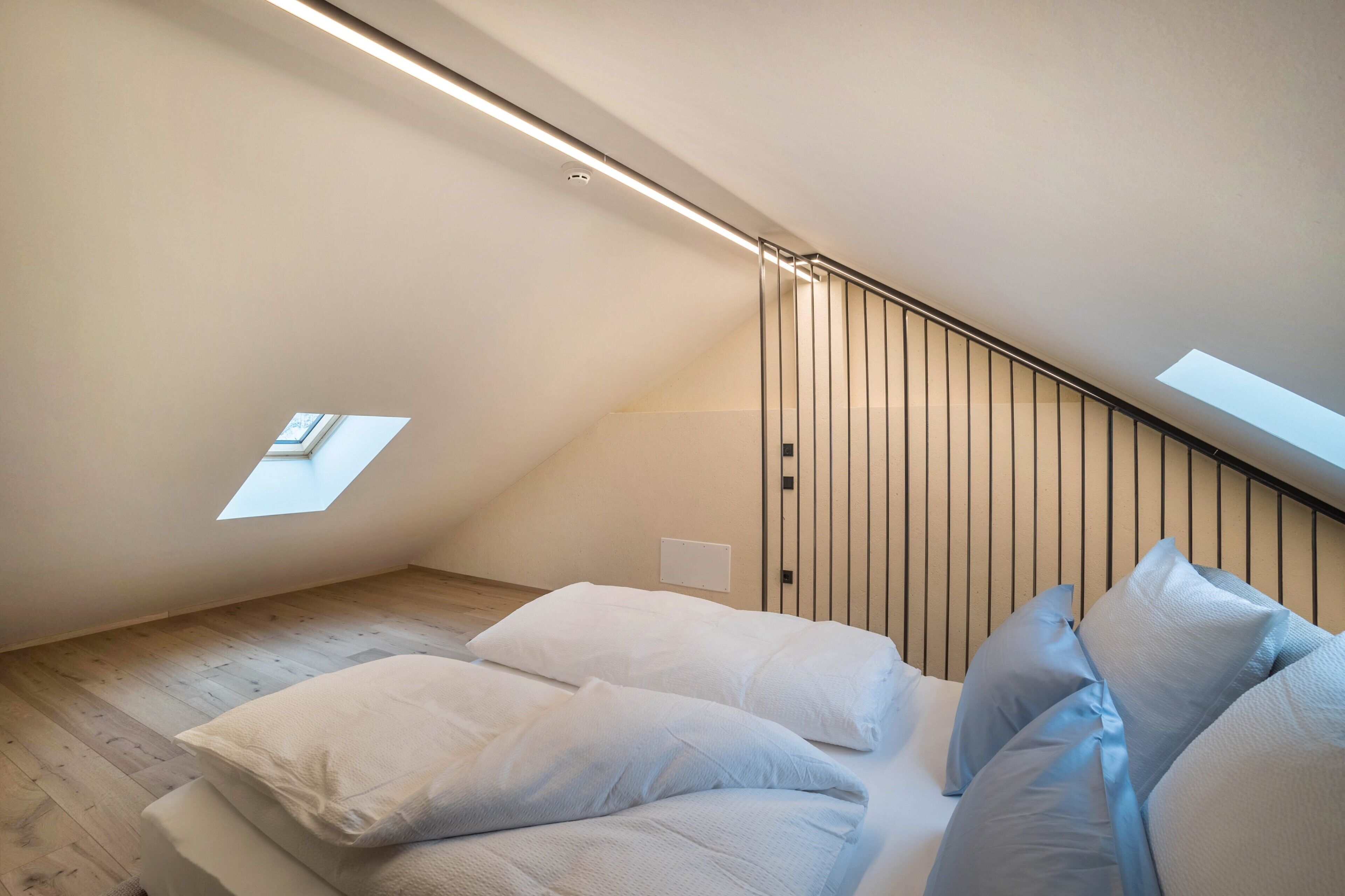 Un dormitorio moderno con techo inclinado, tragaluces y cama con almohadones blancos y azules.