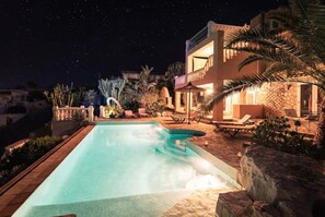 Un área de piscina nocturna con piscina cristalina, sillas de descanso y un edificio con balcón.