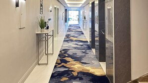 Un pasillo moderno de hotel con alfombra estampada, iluminación empotrada y vista al área de las habitaciones.