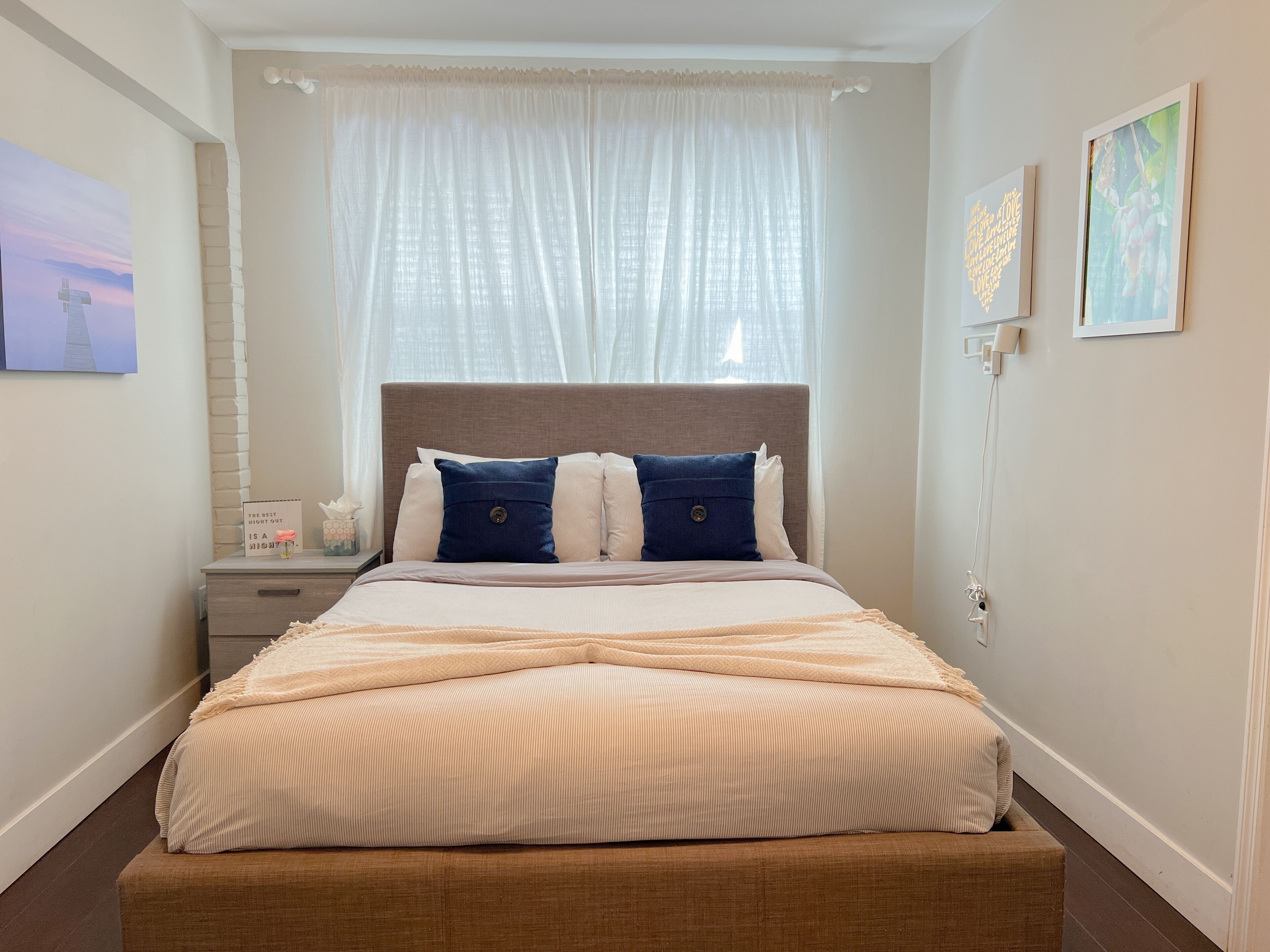 Quarto bem arrumado com cama, mesa de cabeceira e janela com cortinas leves.