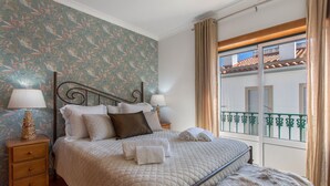 Ein ordentlich bezogenes Bett mit Kissen und Decke, eine Nachttischlampe, ein Nachttisch aus Holz und ein Fenster mit Blick auf Gebäude.
