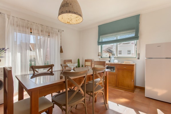 Un comedor con una mesa y sillas de madera, una lámpara colgante, un ventanal con vistas y una zona de cocina con refrigerador.