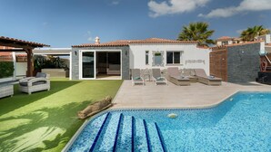 Une piscine avec un bord carrelé, un patio couvert doté de chaises longues et un bâtiment surmonté d’un toit en tuiles.
