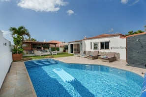 Una piscina con una tumbona flotante, un patio con mobiliario exterior y una casa con techo de tejas.